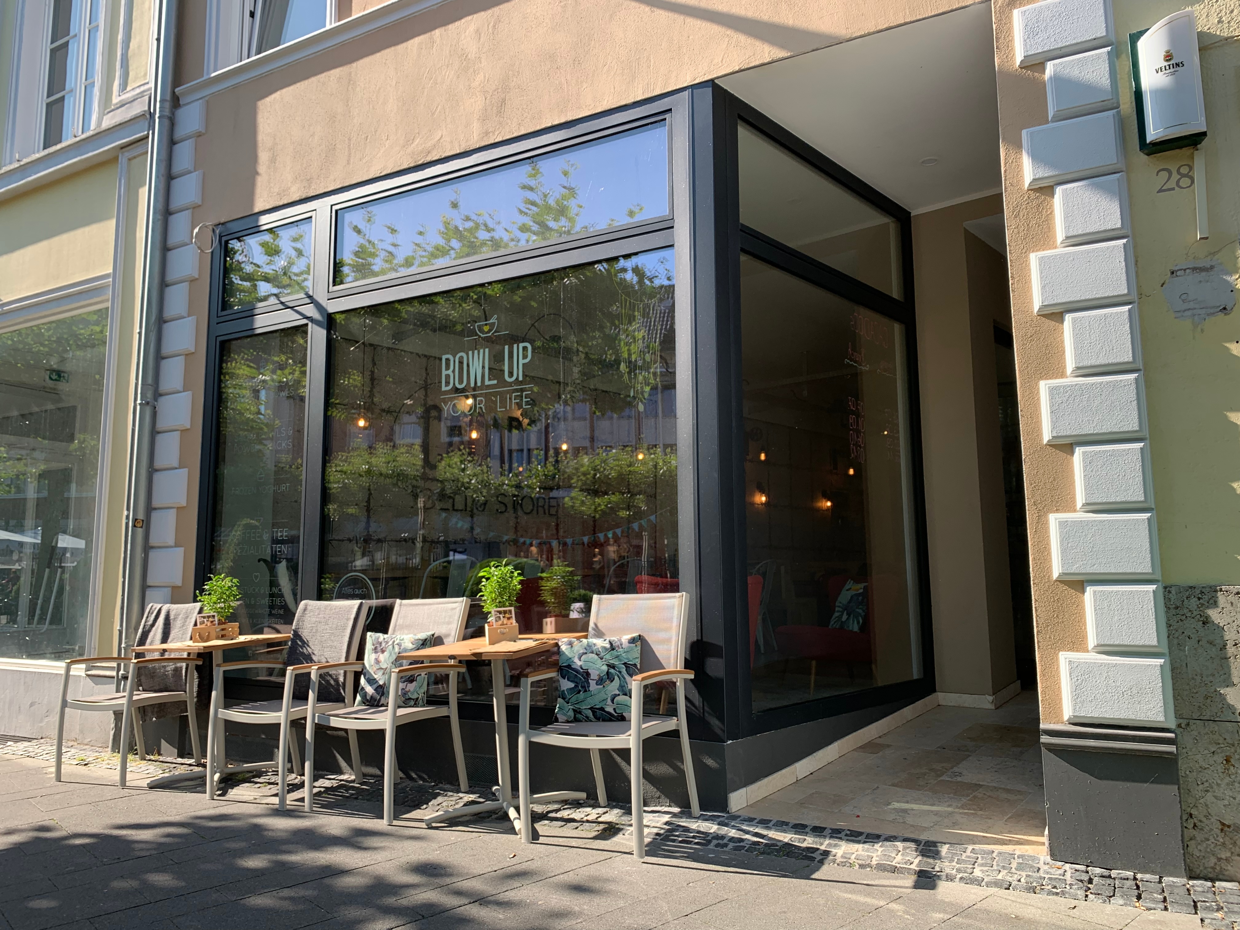 Holz-Aluminium Fenster in einem Straßencafe an welchem Stühle stehen. Rechts ist der Durchgang zur integrierten Haustür