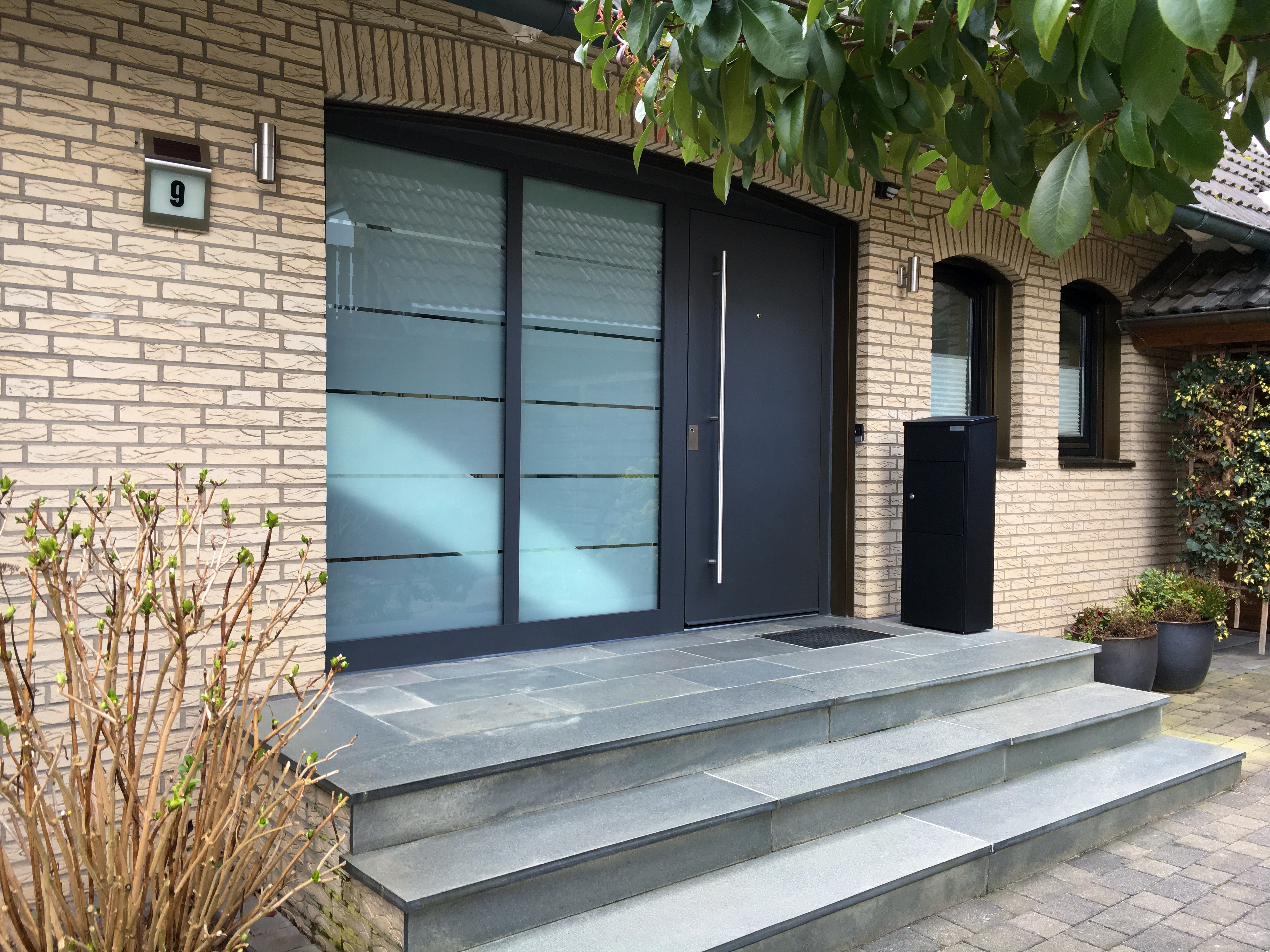 Außenbereich einer schwarzen Haustüre mit doppeltem Glas-Seitenteil an der linken Seite. Der Einang befindet sich oberhalb dreier Treppenstufen. Rechts ist ein schwarzer Säulen-Briefkasten